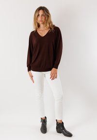 Maglione marrone a V con maniche lunghe, abbinato a pantaloni bianchi aderenti e stivali neri alla caviglia. Texture liscia, vestibilità rilassata, design minimal.