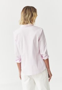 Chemise rose clair à manches longues avec manches retroussées et ourlet arrondi. Le matériau semble lisse ; présente un col classique et un yoke au dos.