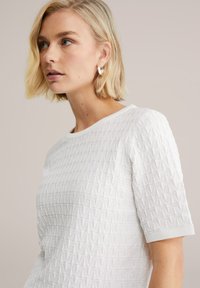 Teksturowany biały top z krótkim rękawem, zdobiony geometrycznym wzorem i prążkowanym kołnierzem. Modelka ma krótkie blond włosy i nosi srebrną kolczyk w kształcie koła.