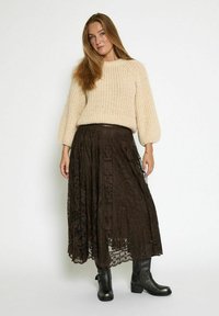 Beige strikket sweater med pufærmer, parret med en lang, mørkebrun blondejakke og sorte ankelstøvler. Nederdelen har intrikate mønstre.