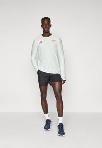 Hellgrünes langärmliges Sportshirt mit pinkem Logo, kombiniert mit schwarzen Shorts und dunkelblauen Laufschuhen. Schlichter Hintergrund ohne zusätzliche Details.