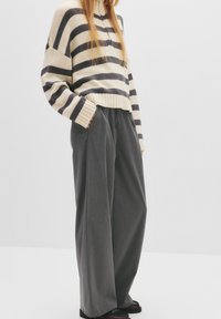 Kvinde iført en cremet og sort stribet sweater kombineret med løse grå brede bukser og mørke sko, stående foran en ensfarvet baggrund.