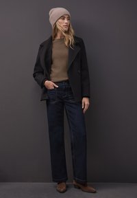 Manteau noir croisé porté sur un pull beige, associé à un jean large bleu foncé et des bottines marron. Bonnet gris clair sur la tête.