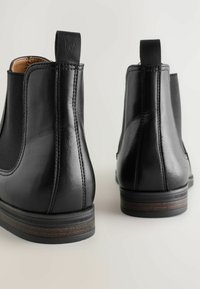 Botas de tobillo de cuero negro con paneles laterales elásticos, textura suave, punta redonda y un detalle de diseño minimalista en la suela.