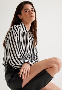 Camisa de manga larga a rayas blancas y negras con cuello abotonado, combinada con una falda de cuero negra. Tela suave y ajuste relajado.
