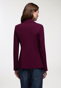 Blazer bordeaux con un design aderente, maniche lunghe e colletto a punta. Tessuto morbido con orlo leggermente svasato. Vista posteriore mostrata.