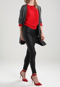 Vrouw in een grijze cardigan, rode geribbelde trui, zwarte leren broek en rode hoge hakken met enkelbandjes.
