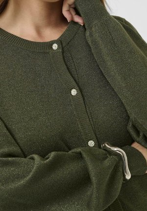 Gros plan d'une personne portant un cardigan en tricot vert olive avec des boutons décoratifs et un bracelet argenté ondulé au poignet.