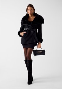 Zwarte jas met nepbontkraag, getailleerde taille en zachte textuur. Gecombineerd met een kleine zwarte handtas met gouden accenten en laarzen met hoge hakken.