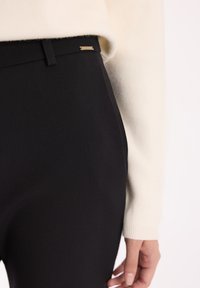 Czarne spodnie w zbliżeniu, charakteryzujące się gładką teksturą, dopasowanym krojem i małym metalowym akcentem na pasku, w połączeniu z swetrem w kolorze kremowym.