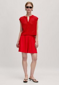Rotes gestricktes ärmelloses Top mit Knopfverschluss und offenem Webdesign, kombiniert mit passenden roten Shorts. Das Modell trägt schwarze Sonnenbrillen und Sandalen.