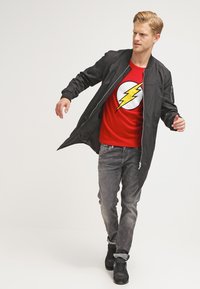 Schwarze Bomberjacke über einem roten T-Shirt mit einem Blitzlogo, kombiniert mit grauen Jeans und schwarzen Sneakers. Insgesamt leichte Textur.