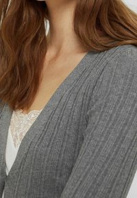 Grå ribbade koftan med en djup V-ringning och texturerad tyg, lagd över en vit blus med spetskant.