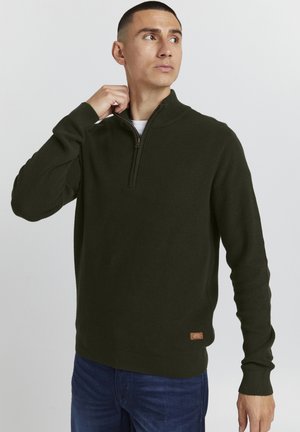 Dark green ribbed sweater with a zip collar, featuring long sleeves and a brown label at the hem. Worn with blue jeans.