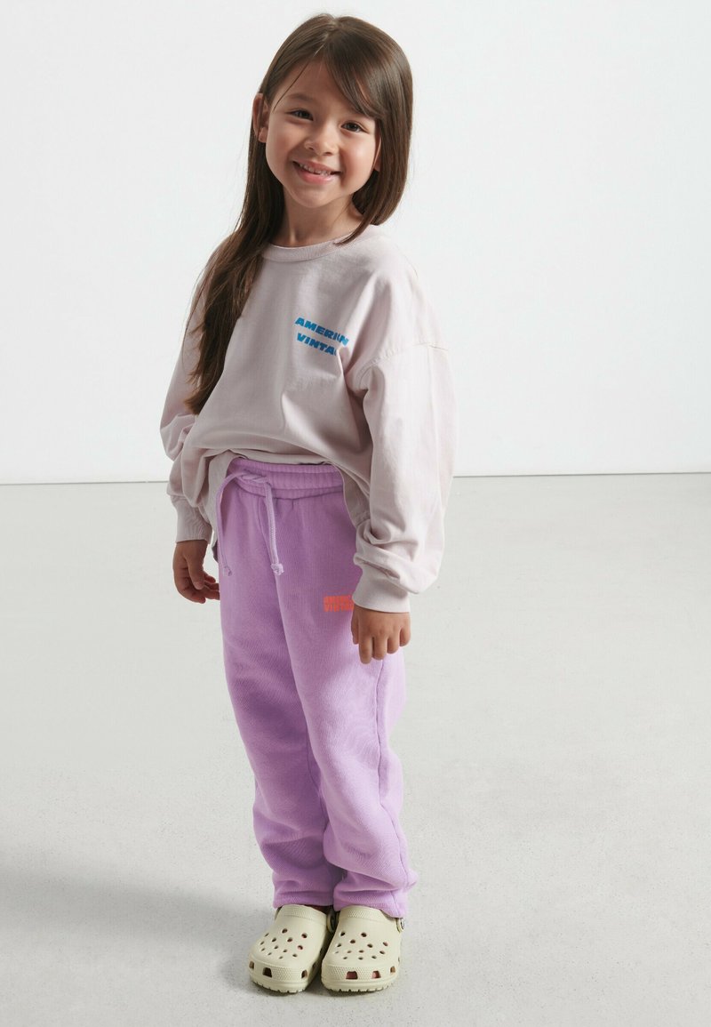 Light pink long-sleeve shirt with blue text, paired with loose purple sweatpants featuring orange text, and green clogs.