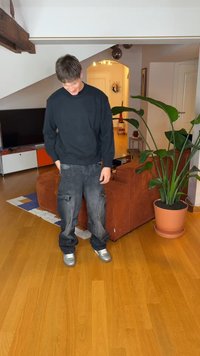 Svart genser, vide grå cargobukser med lommer og sølvfargede joggesko. Innendørs setting med tregulv og potteplante.