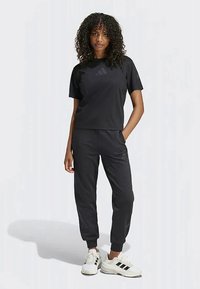 Görbe hajú nő fekete Adidas pólót, fekete joggert és fekete csíkos fehér Adidas sportcipőt visel.