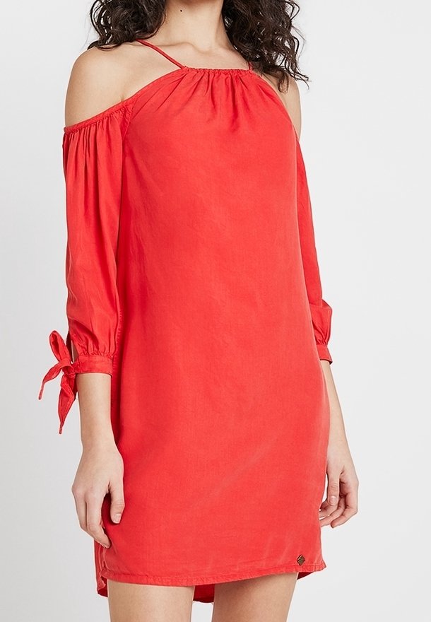 Femme portant une robe rouge à épaules dénudées avec des manches trois-quarts attachées aux poignets, debout devant un fond blanc uni.