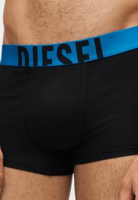 Gros plan d'une personne portant un boxer noir avec une ceinture bleue portant le mot "DIESEL" en lettres noires.