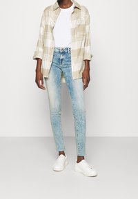 Camisa de cuadros beige y blanca, camiseta blanca, jeans ajustados azul claro y zapatillas blancas. La camisa tiene cierre de botones y mangas dobladas.