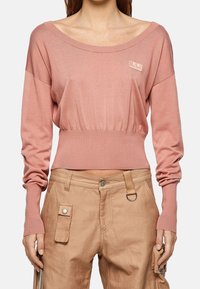 Jasnoróżowy, długi sweter z długim rękawem, szerokim dekoltem, marszczoną talią i subtelnym detalem z logo. W połączeniu z beżowymi spodniami typu cargo z kieszeniami.