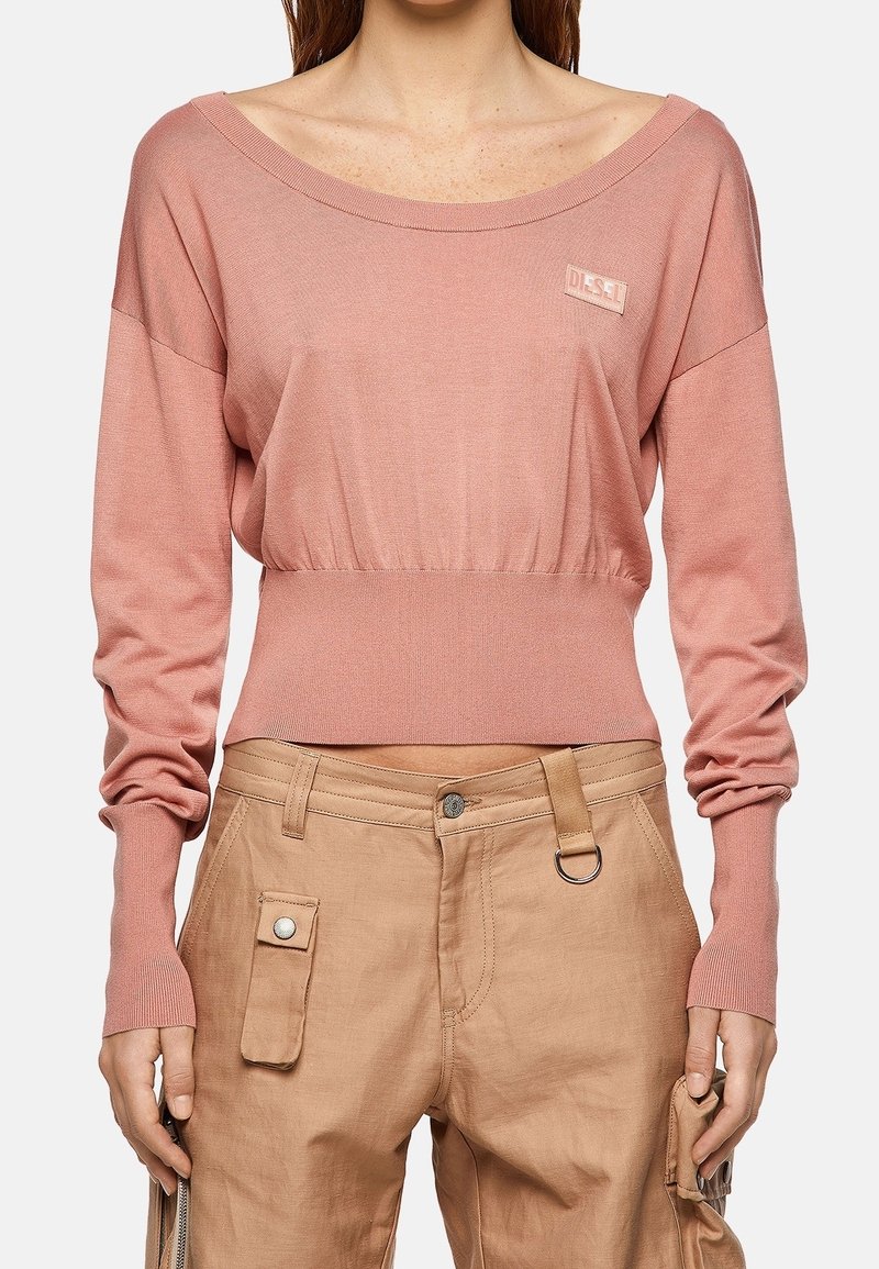 Jasnoróżowy, długi sweter z długim rękawem, szerokim dekoltem, marszczoną talią i subtelnym detalem z logo. W połączeniu z beżowymi spodniami typu cargo z kieszeniami.