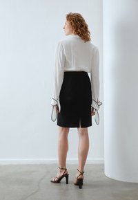 Biela blúzka s vypchatými rukávmi a čierna sukňa s rozparkom na zadnej strane. Čierne sandále na vysokých opätkoch dopĺňajú outfit. Hladké textúry.