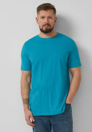 Baarddragende man met kort haar, gekleed in een felblauw shirt met korte mouwen en blauwe spijkerbroek, staand tegen een lichtgrijze muur.