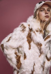 Fuskpälsjacka i vitt med bruna fläckar, med lös passform, hög krage och texturerad yta. Diskret design utan synliga detaljer.