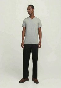 Tall man standing wearing a light gray short-sleeve V-neck shirt, black wide-leg pants, and brown shoes against a plain background.