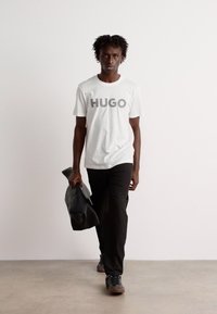 Hvit t-skjorte med grå "HUGO"-logo, svarte bukser og mørke sko. Modellen holder en svart skulderveske og står mot en nøytral bakgrunn.