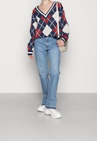 Pull en maille à col en V avec motif à losanges rouges, bleu marine et blancs ; associé à un jean bleu et des baskets blanches épaisses ; petit sac beige.