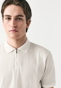 Polo beige clair avec un motif à carreaux texturé, doté d'un col zippé et de manches courtes, fabriqué en tissu respirant.