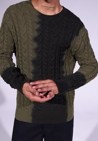 Homme portant un pull en tricot torsadé vert et noir, les mains jointes, avec un tatouage « crois en toi » visible sur le cou.