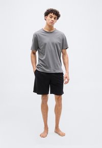 Νεαρός άνδρας όρθιος, ξυπόλυτος, φορώντας γκρι μπλούζα και μαύρο σορτς, μπροστά σε απλό ανοιχτό γκρι φόντο.