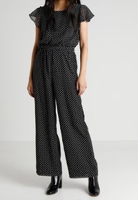 Combinaison noire à pois avec manches courtes transparentes, coupe à jambes larges et taille cintrée. Présente une texture lisse et un tissu léger.