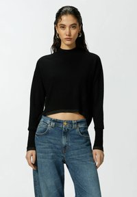 Crna skraćena džemper sa visokim izrezom i rebrastim manžetama, u kombinaciji sa širokim svijetloplavim farmerkama s prednjim džepovima i mesinganim detaljima.