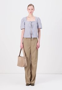 Femme portant un haut à rayures à manches courtes, un pantalon large beige, tenant un sac fourre-tout beige, se tenant devant un fond blanc uni.