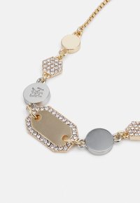 Collana a catena color oro e argento con anelli esagonali e circolari, impreziosita da strass trasparenti e un'etichetta centrale ottagonale dorata.