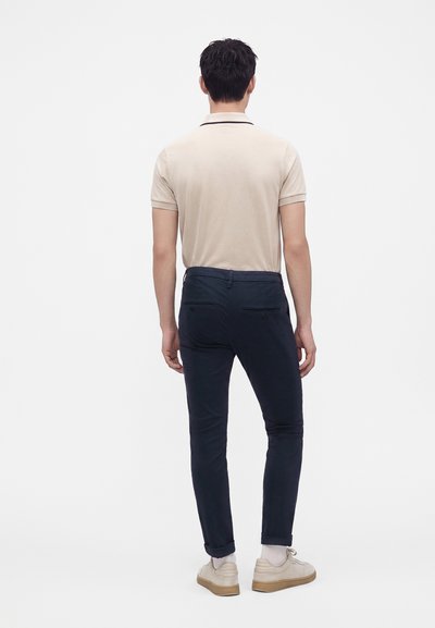 Homme portant un polo beige, un pantalon bleu marine foncé et des baskets beige clair, de dos sur fond blanc uni.