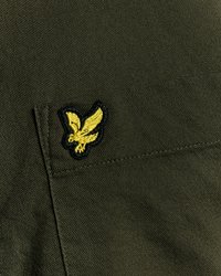 Camisa de tela verde oliva con un bolsillo en el pecho y un logotipo de águila bordado en amarillo con contorno negro. Material texturizado.
