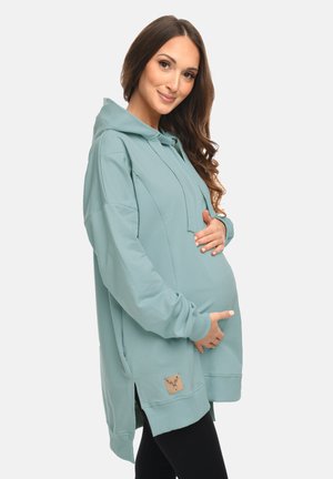 Schwangere Frau in übergroßem türkisfarbenem Hoodie, die sanft ihren Bauch hält und lächelt, vor schlichtem hellem Hintergrund.