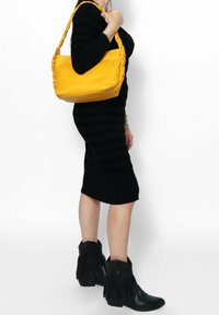 Borsa a tracolla in pelle gialla con manico intrecciato; modello in un vestito nero a coste e stivaletti alla caviglia con frange nere.