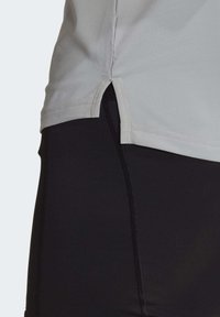 Hellgraues Sportoberteil mit seitlichen Schlitzen, aus glattem Material, kombiniert mit schwarzen Leggings, die eine strukturierte Naht aufweisen.