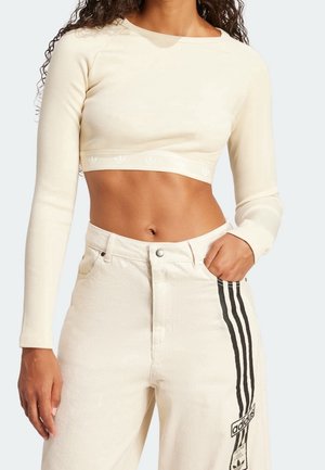 Kobieta w kremowym, długim rękawem, krótkim topie oraz kremowych spodniach Adidas z czarnymi paskami i logo na lewej nodze.