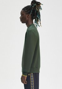 Felpa verde con scollatura rotonda e polsini a costine, abbinata a pantaloni scuri con un motivo a logo dorato sul lato.