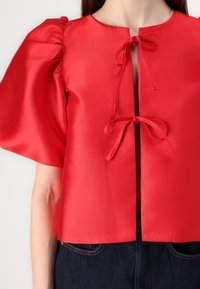Top in raso rosso con maniche a palloncino, scollatura rotonda e legature frontali. Il tessuto ha una superficie liscia con una leggera lucentezza.