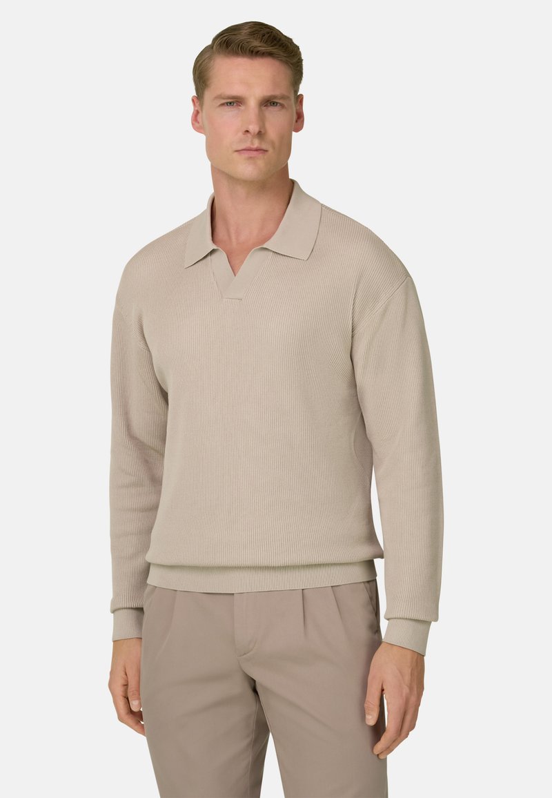 Homme portant un polo en maille à manches longues beige et un pantalon assorti, debout avec une expression neutre devant un fond gris clair.