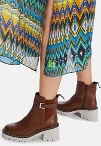 Braune Leder-Ankle-Boots mit strukturierter Oberfläche, grauen Blockabsätzen, Reißverschluss und seitlichem Elastik. Mit einem metallischen Schnallen-Detail akzentuiert.