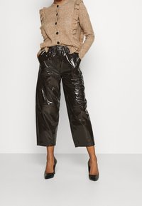 Cardigan marron froncé avec des boutons noirs, associé à un pantalon noir cassé à taille haute et brillant. Des escarpins noirs à talon aiguille complètent le look.